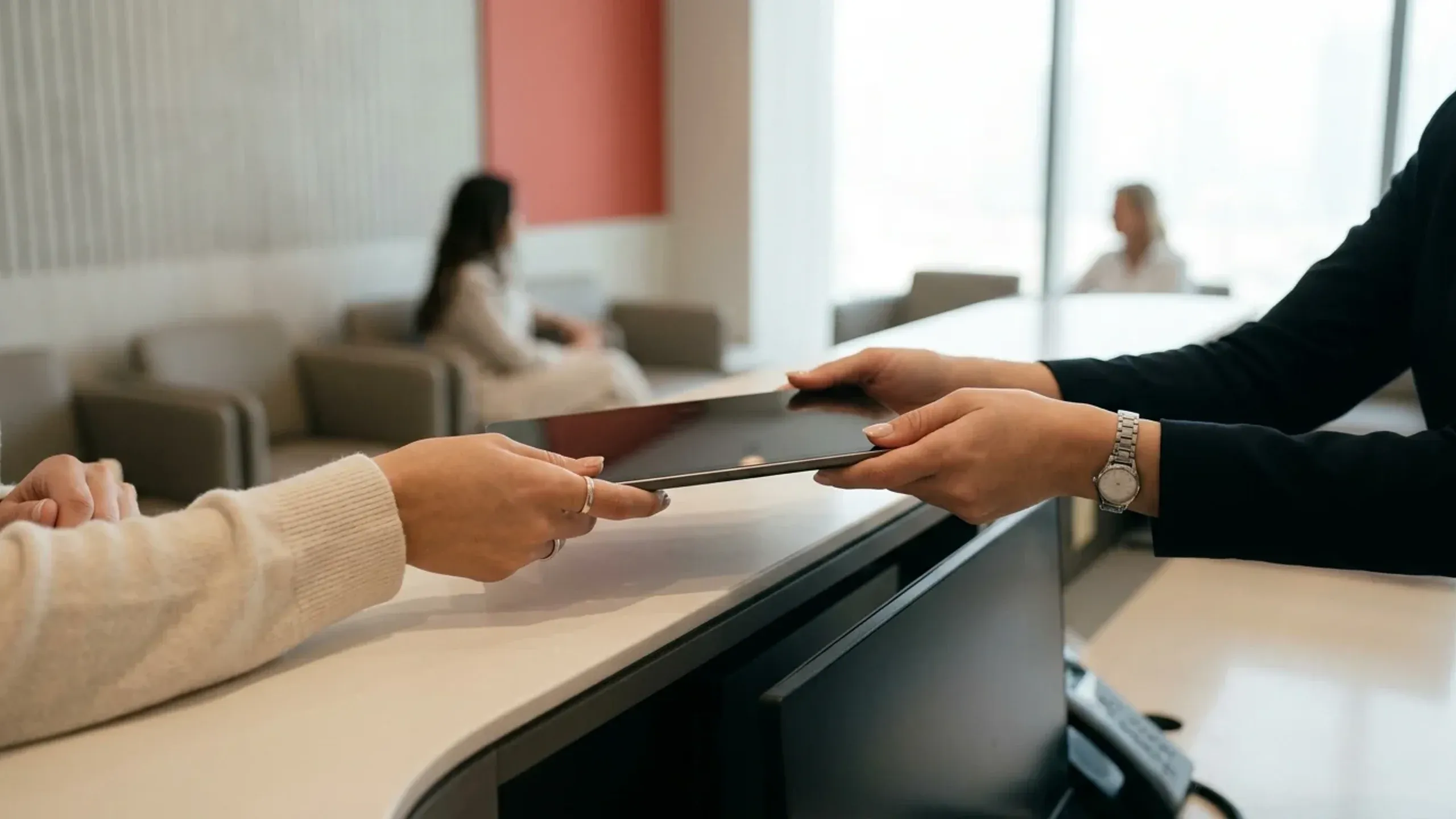 Two pairs of hands passing a closed tablet across a clinic reception desk